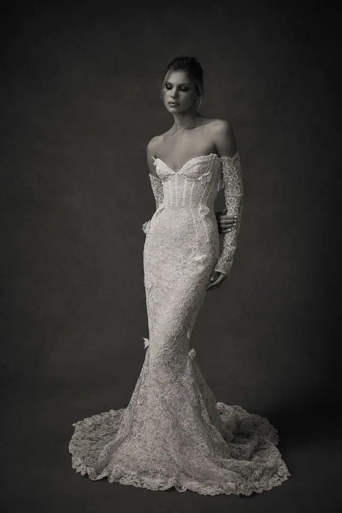 A woman in a form-fitting, lace wedding gown with long gloves, posing against a dark background.