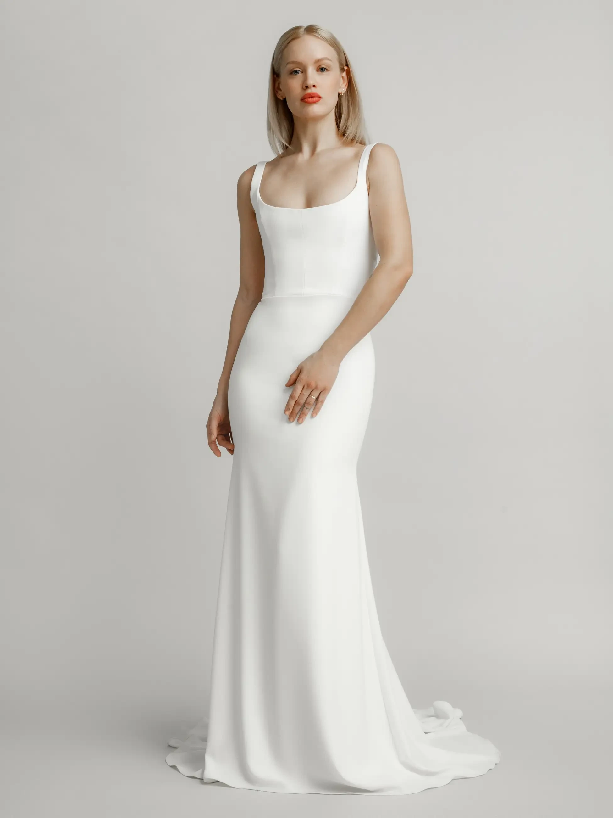 Model is wearing a timeless minimalist mermaid gown with a square scoop neckline, sewn straps, and illusion buttons down the back