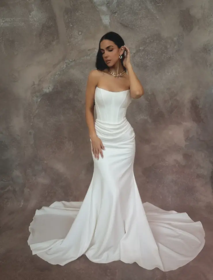 Model is wearing a strapless fitted gown with a structured boned bodice and ruching along the waist and hips