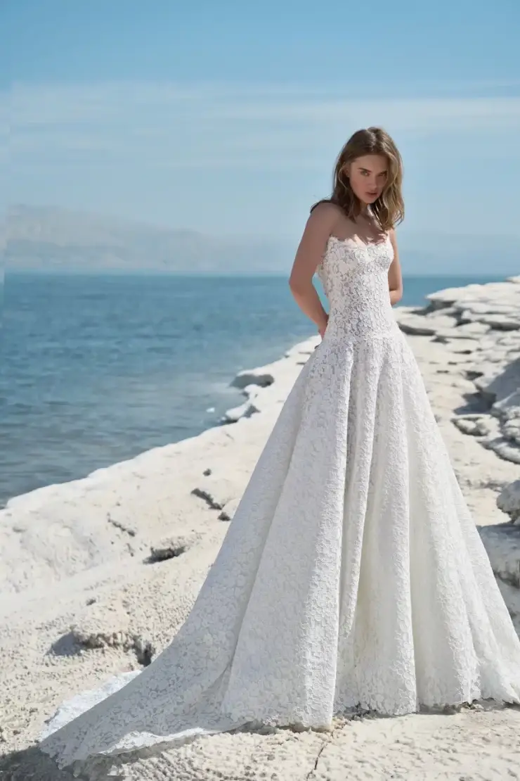 A woman in a white wedding dress stands on a rocky shoreline with the ocean in the background. The dress has intricate detailing and a flowing skirt.