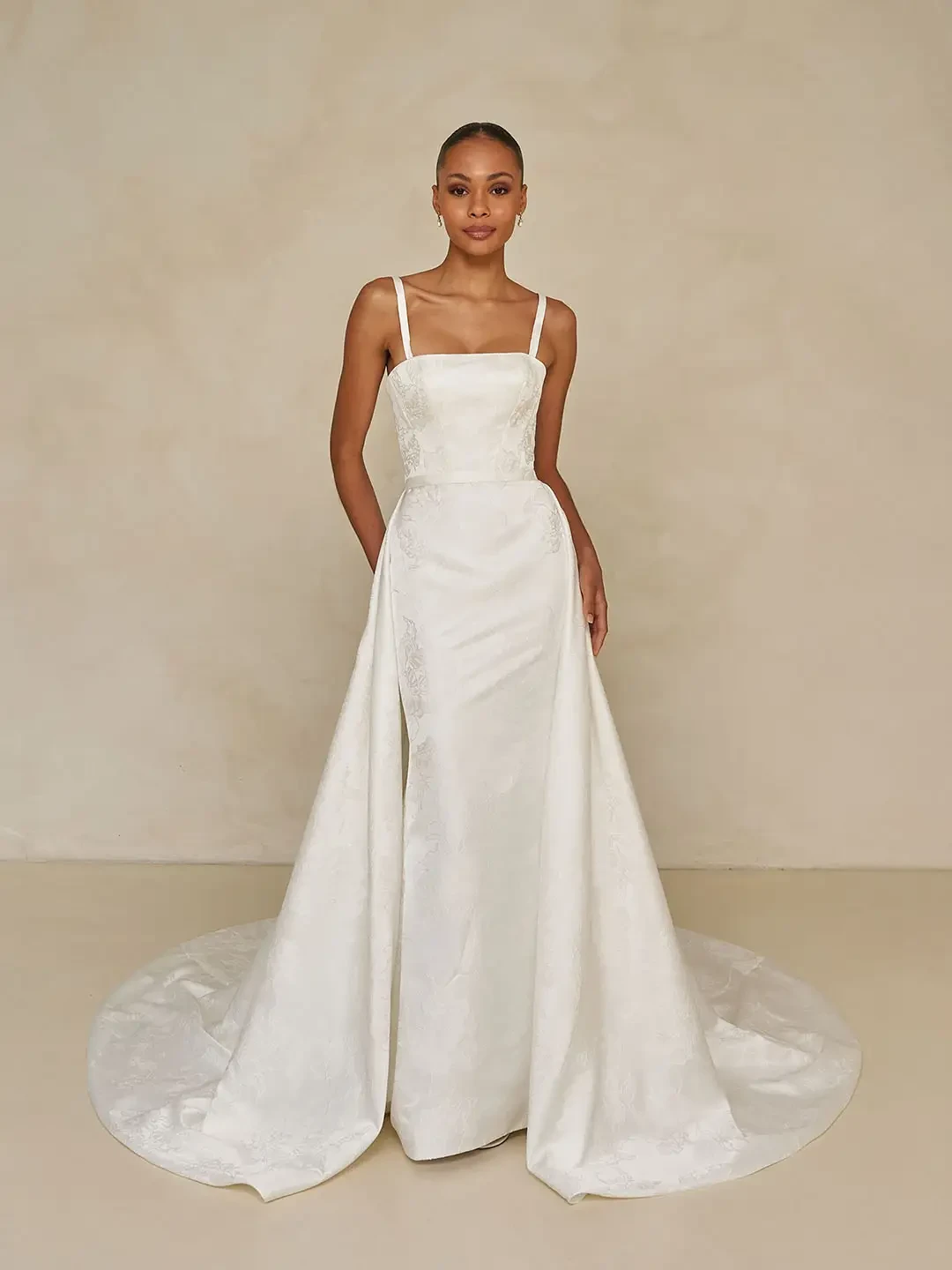 Model is wearing a floral jacquard sheath gown with a square neckline, detachable straps, and an optional A-line overskirt and back bow accessory.