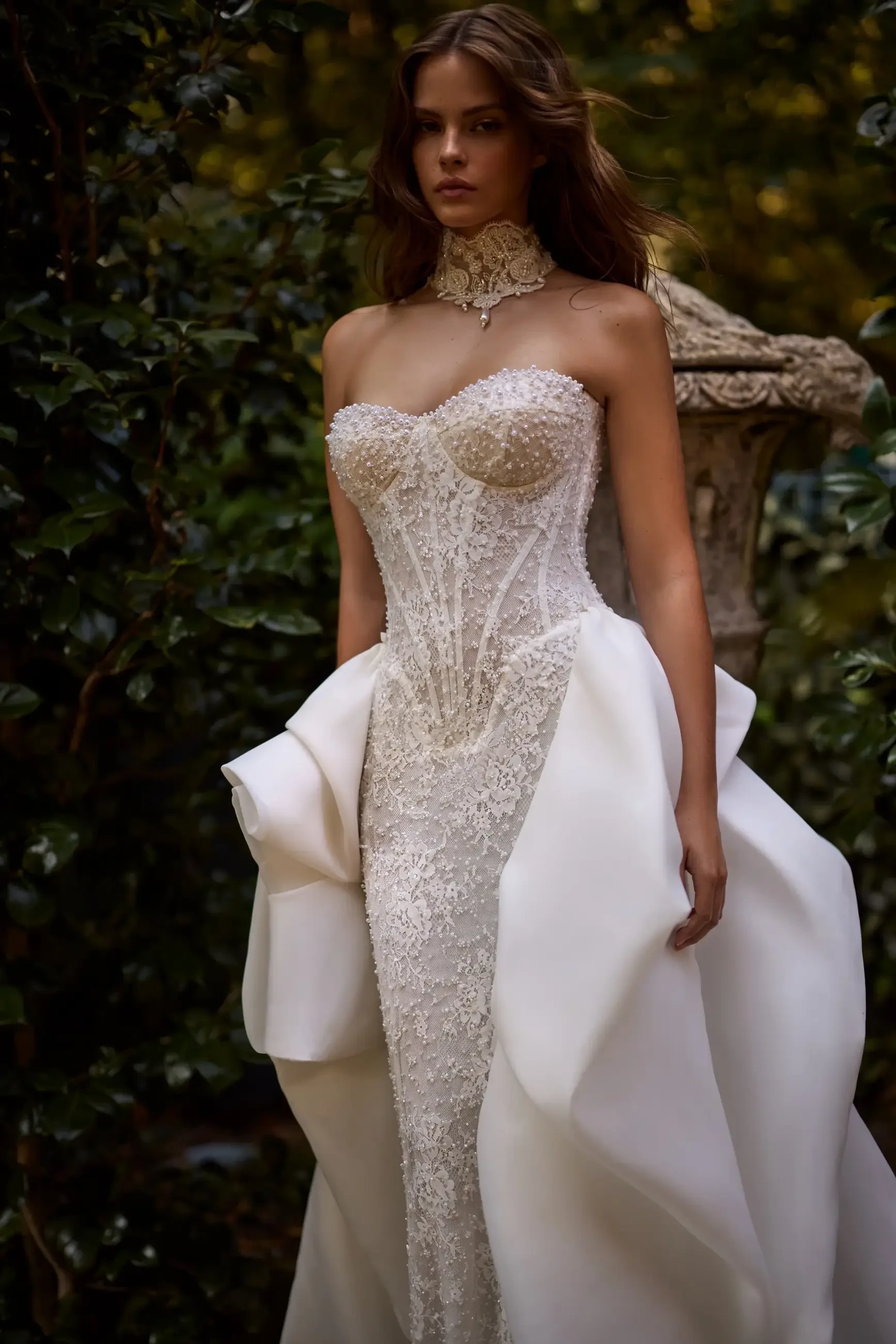 A model wearing an elegant, intricately detailed bridal gown with a fitted bodice and flowing train, walking through a lush outdoor setting.