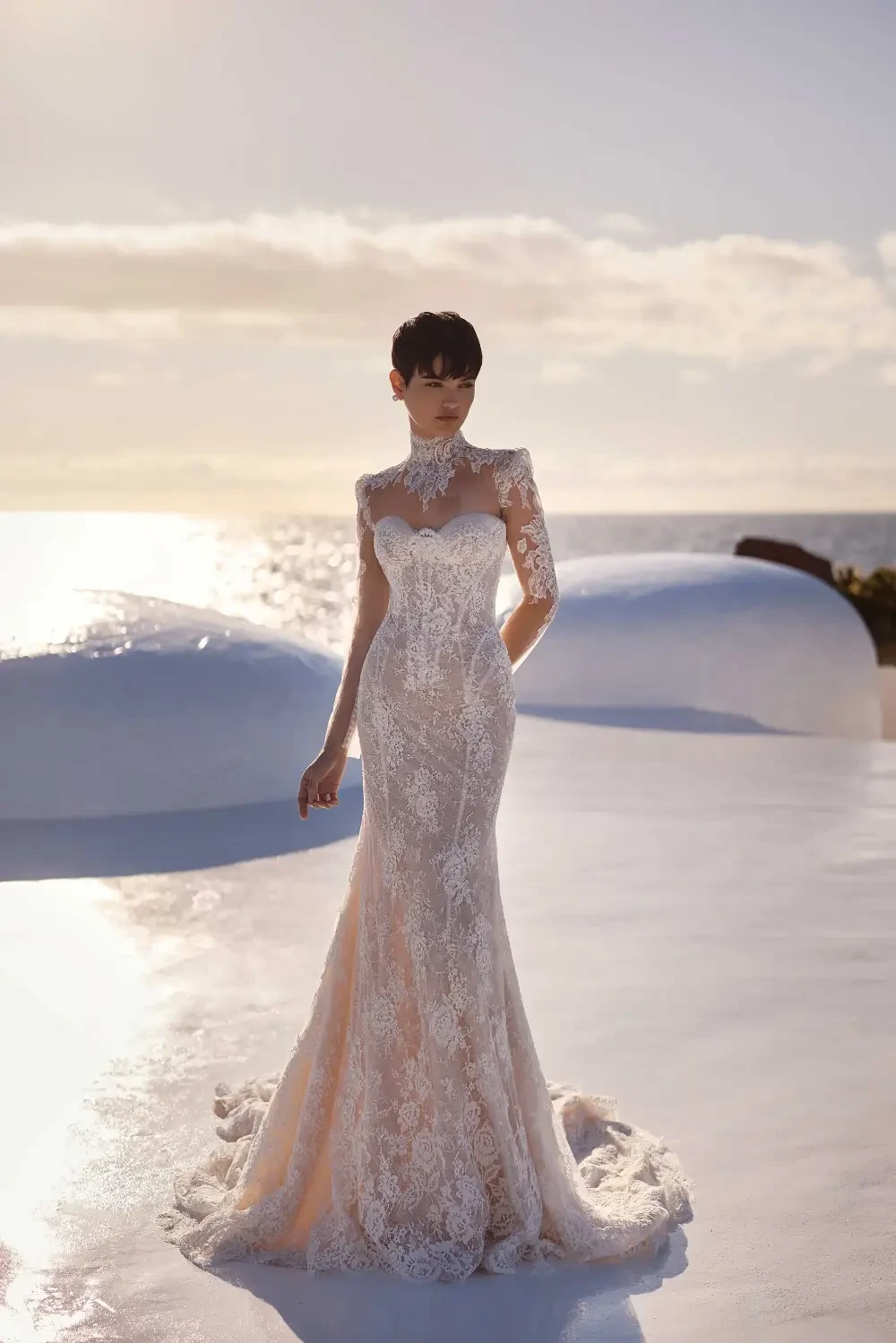 A woman in a beautiful lace wedding dress stands against a scenic backdrop of the ocean and sky, with dramatic lighting highlighting her figure.