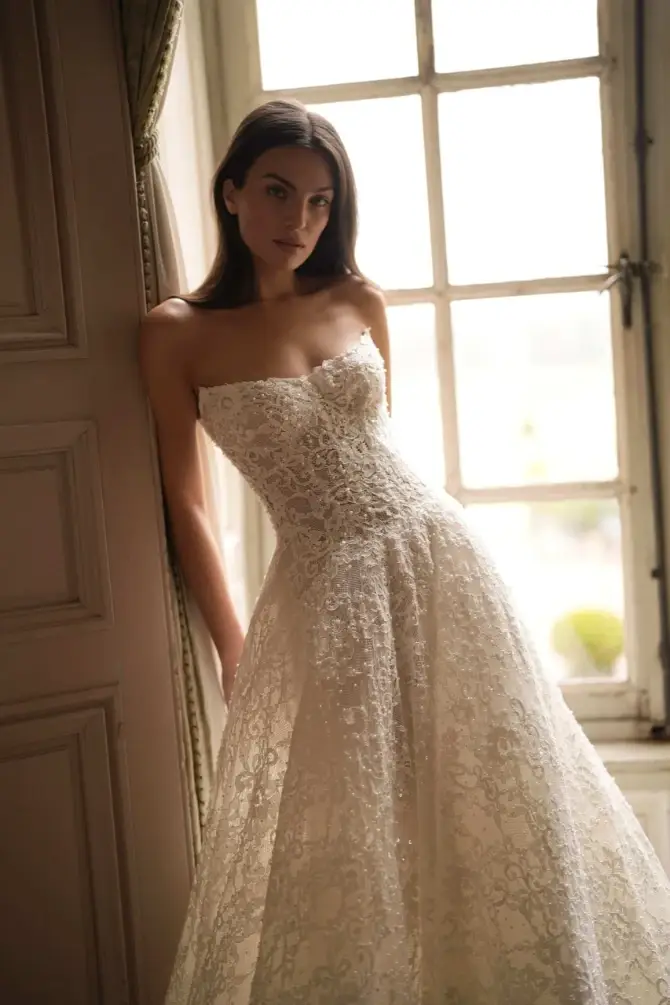 Woman in a strapless lace wedding gown leans against a wall by a sunlit window, creating a serene and elegant atmosphere.