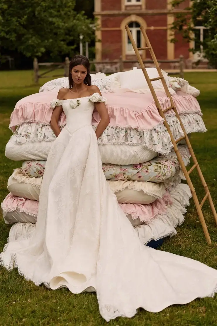 A woman in a long, white off-the-shoulder wedding dress stands next to a large stack of colorful, layered bedding outdoors, with a ladder leaning against the stack and trees in the background.