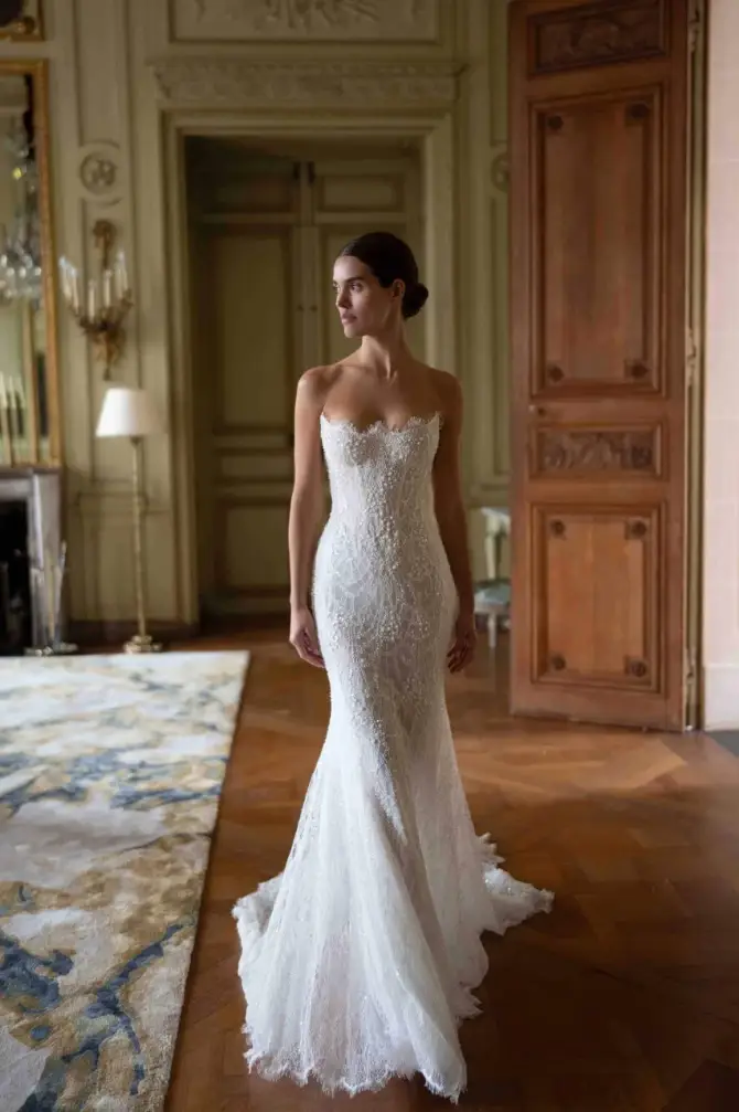 A woman in a lace strapless wedding gown stands on an ornate hardwood floor in an elegant room. Her gaze is serene, conveying a dreamy, romantic mood.