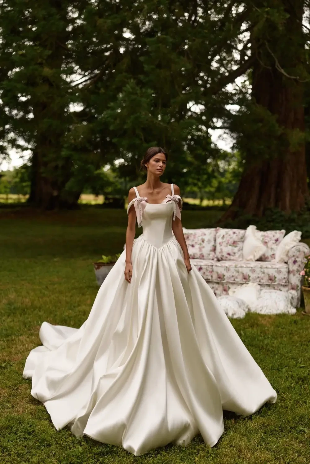 A woman in a flowing white wedding dress stands in a lush outdoor setting with trees in the background.