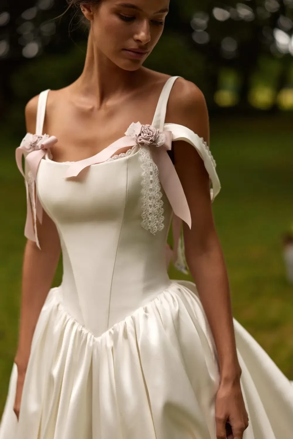 A woman in a flowing white wedding dress stands in a lush outdoor setting with trees in the background.