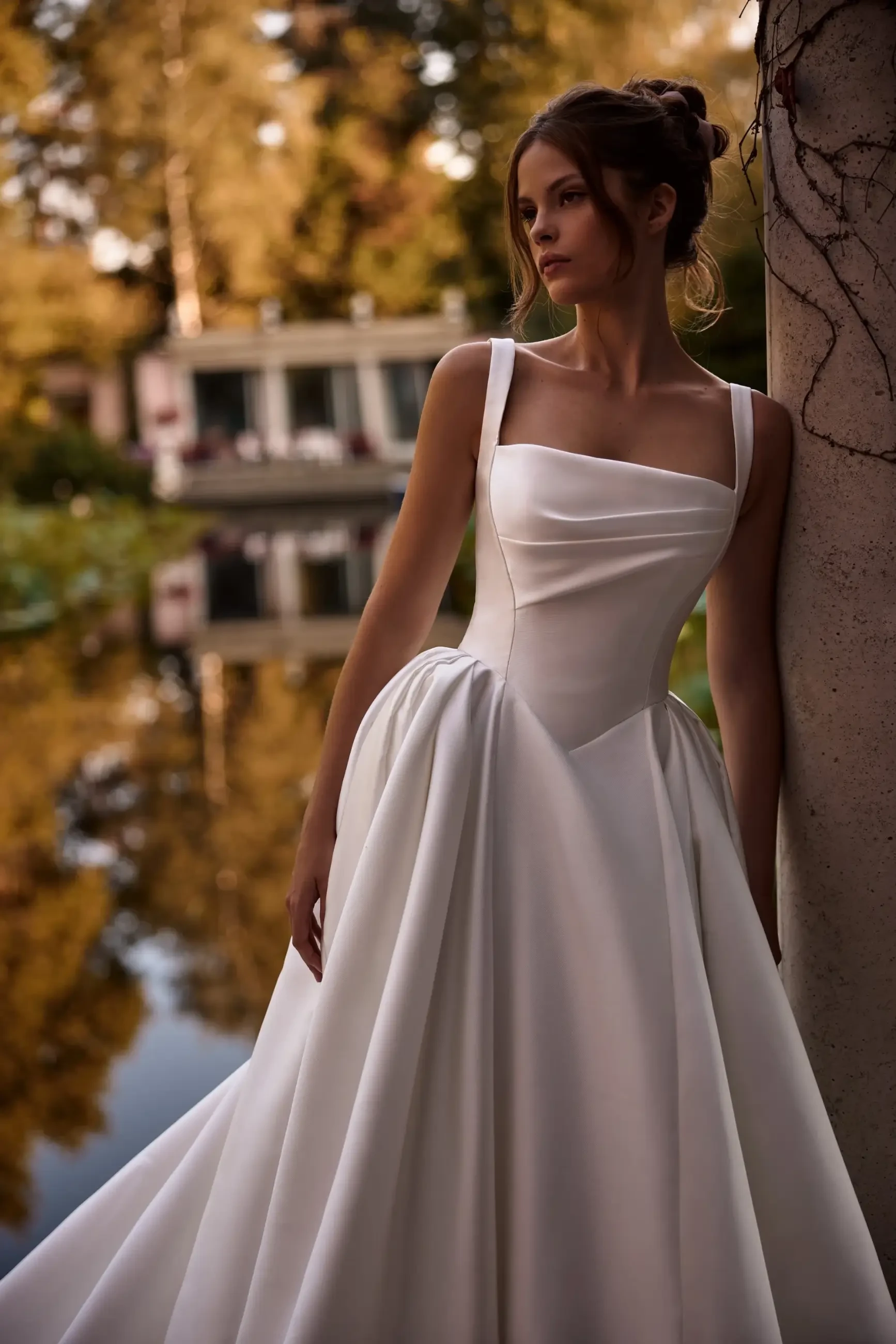 A woman in a white dress stands by a stone pillar, with a reflection of trees and a building in the water behind her.