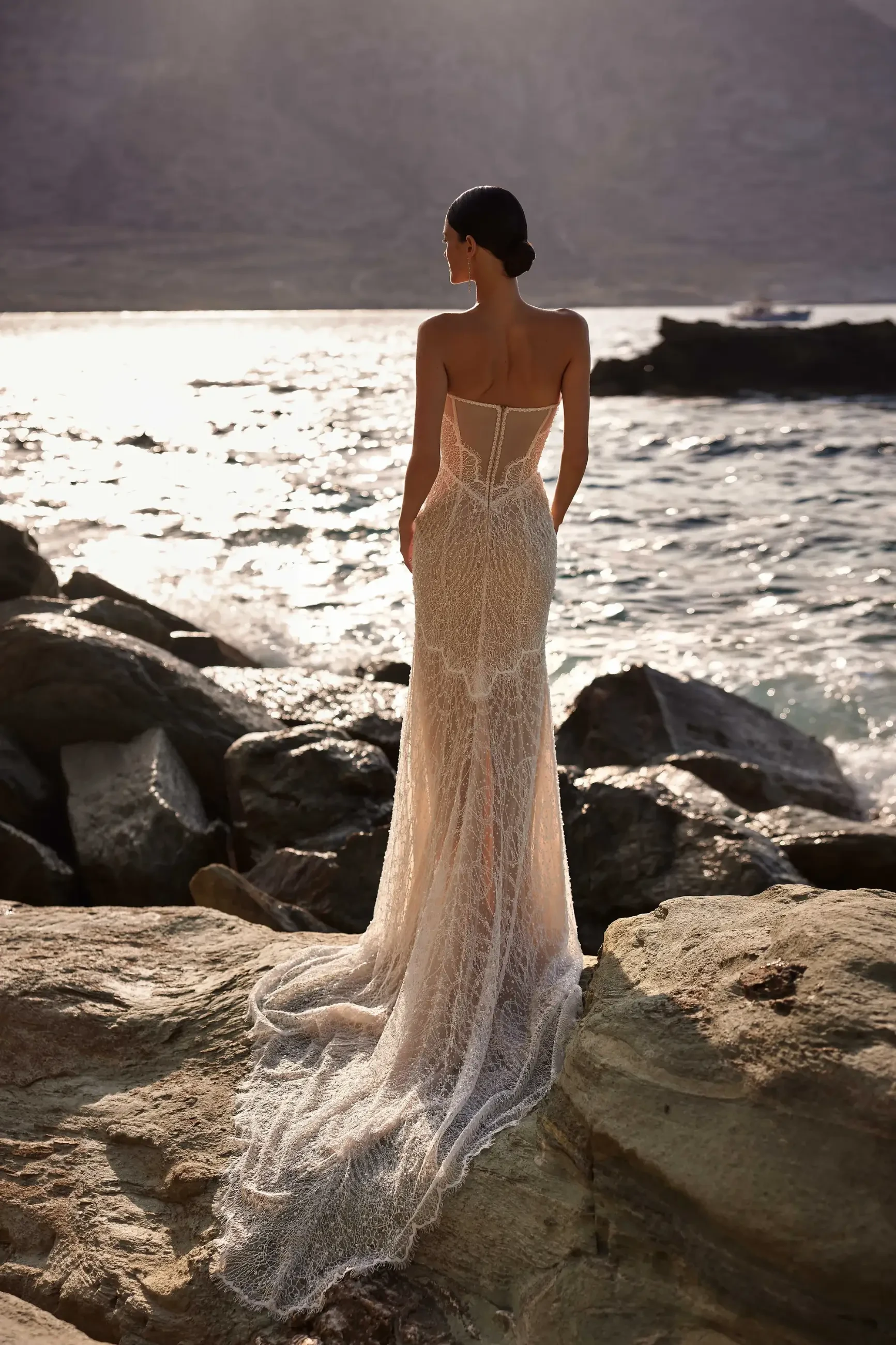 A woman in a flowing, elegant dress stands on rocky terrain by the water, looking out at the ocean. The dress has intricate details and a long train that cascades behind her.