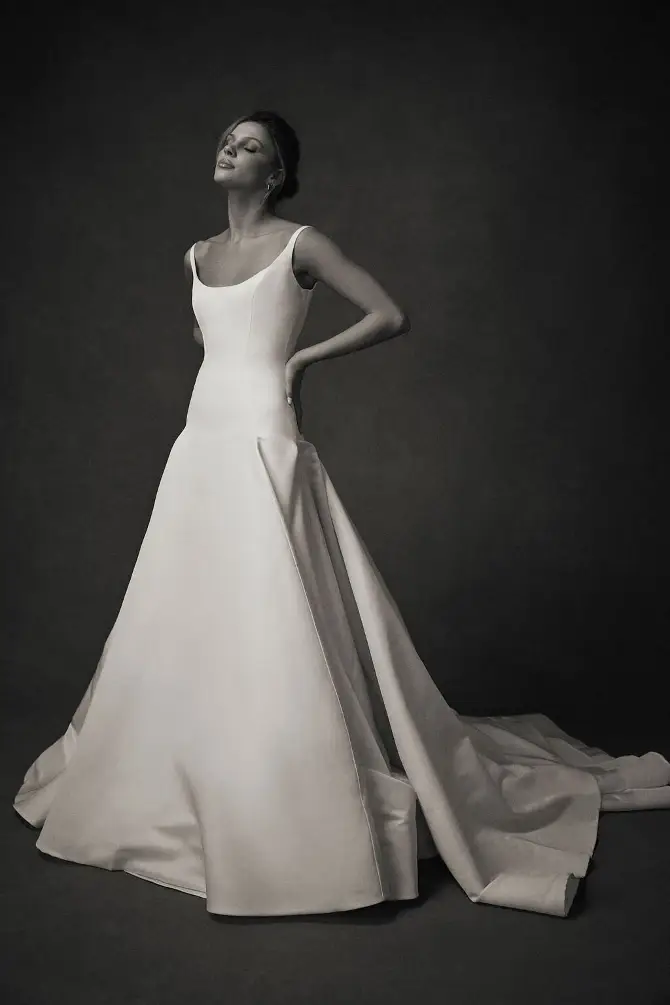 A model poses elegantly in a long, flowy wedding dress with a fitted bodice and wide skirt, set against a dark background.