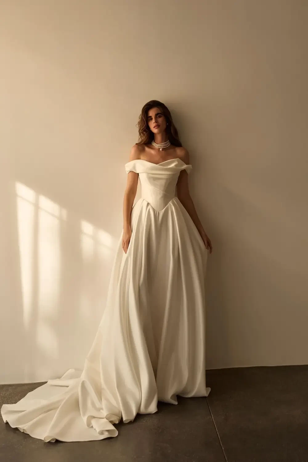 A woman in an elegant white off-shoulder gown stands against a plain wall, with light casting soft shadows.