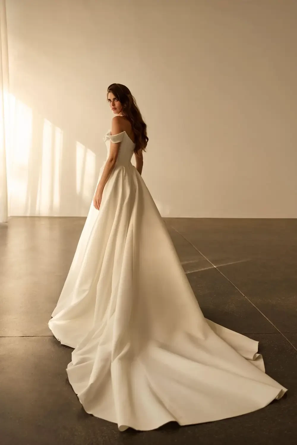 A woman in an elegant white off-shoulder gown stands against a plain wall, with light casting soft shadows.