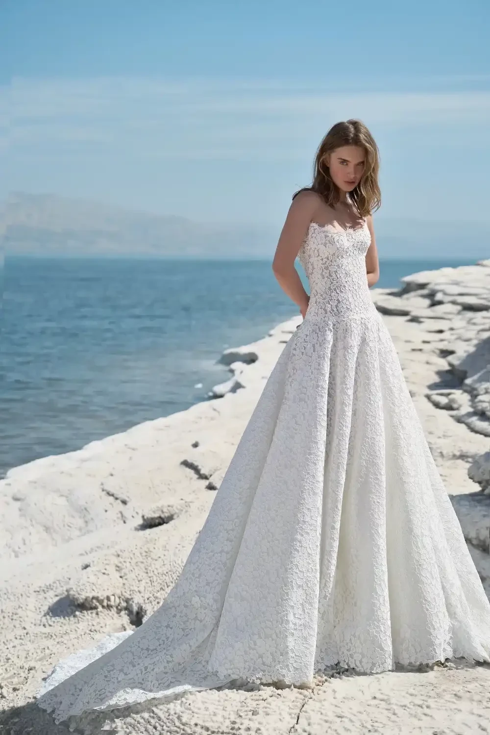 A woman in a stunning white gown stands on a rocky shoreline, overlooking a calm sea.