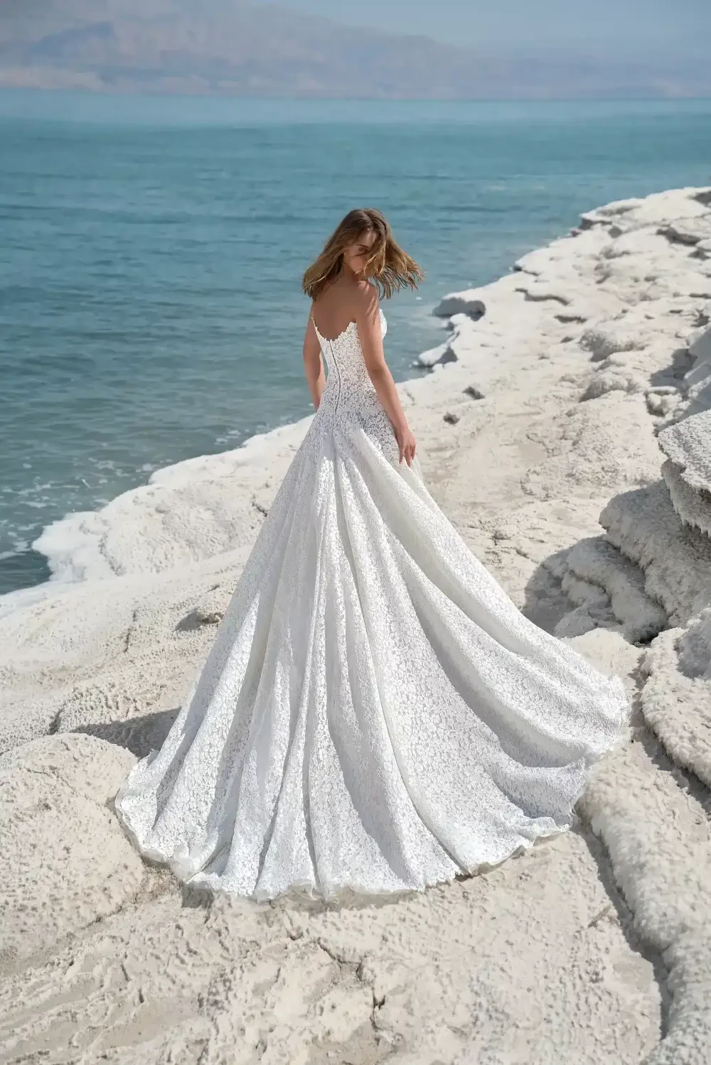 A woman in a stunning white wedding gown stands on a rocky shoreline with the ocean in the background.