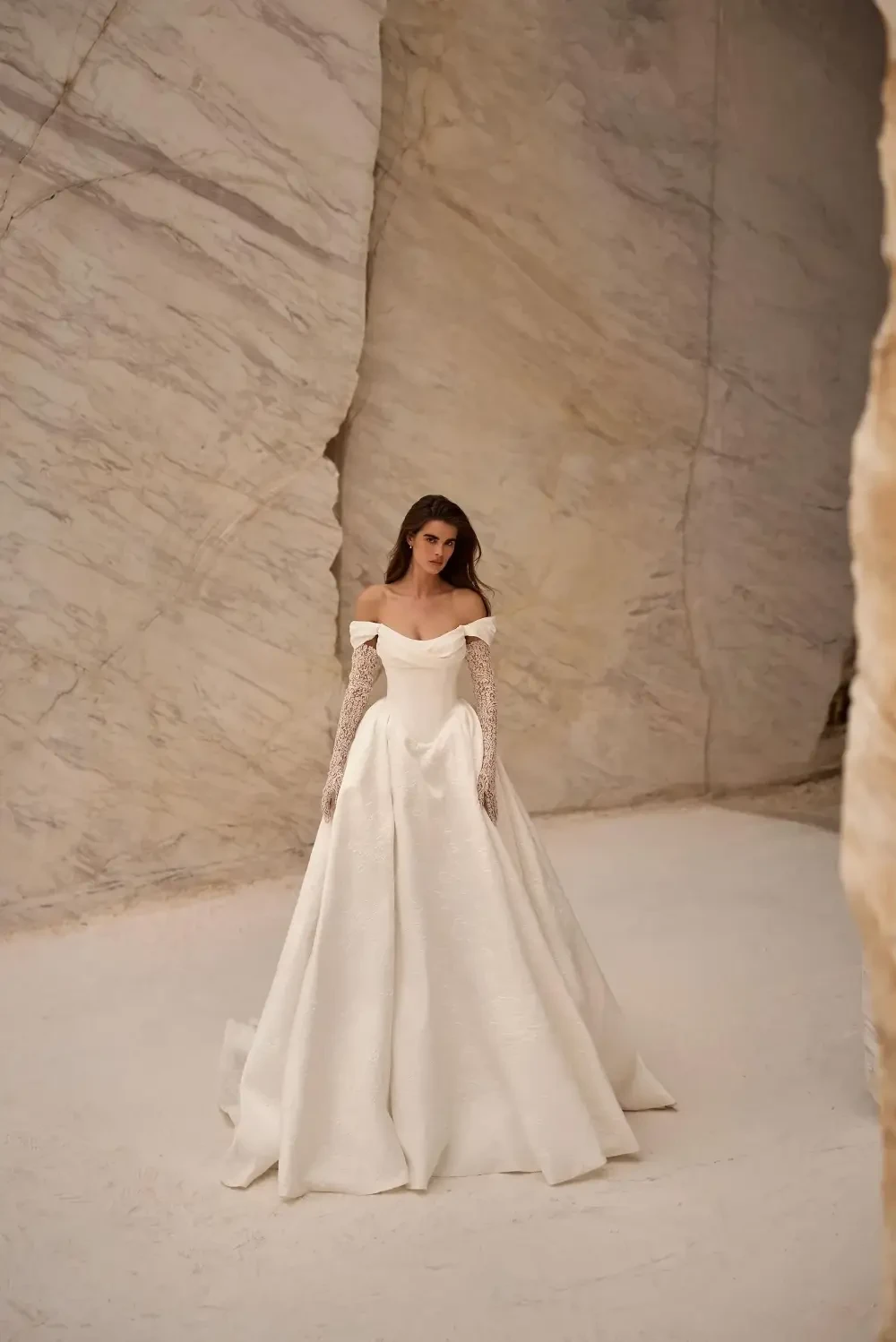 A model wears an elegant off-shoulder white wedding gown with lace sleeves, standing in a minimalistic, marble-like setting.