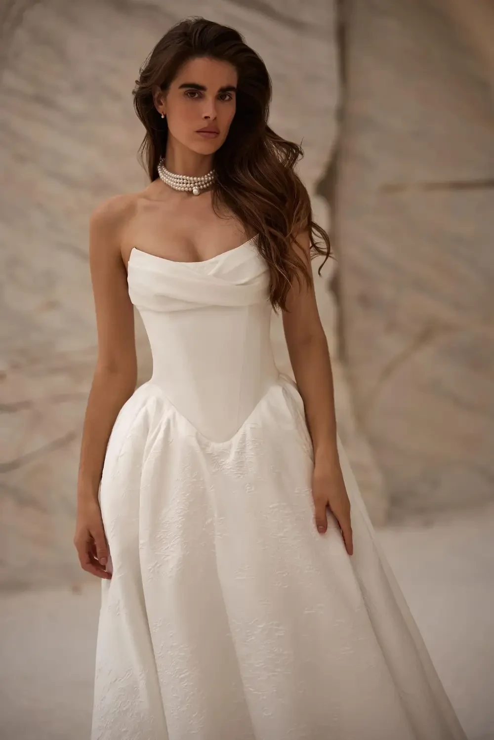 A woman wearing a strapless white wedding dress, with a flowing skirt and a draped bodice, posing confidently in a serene setting.