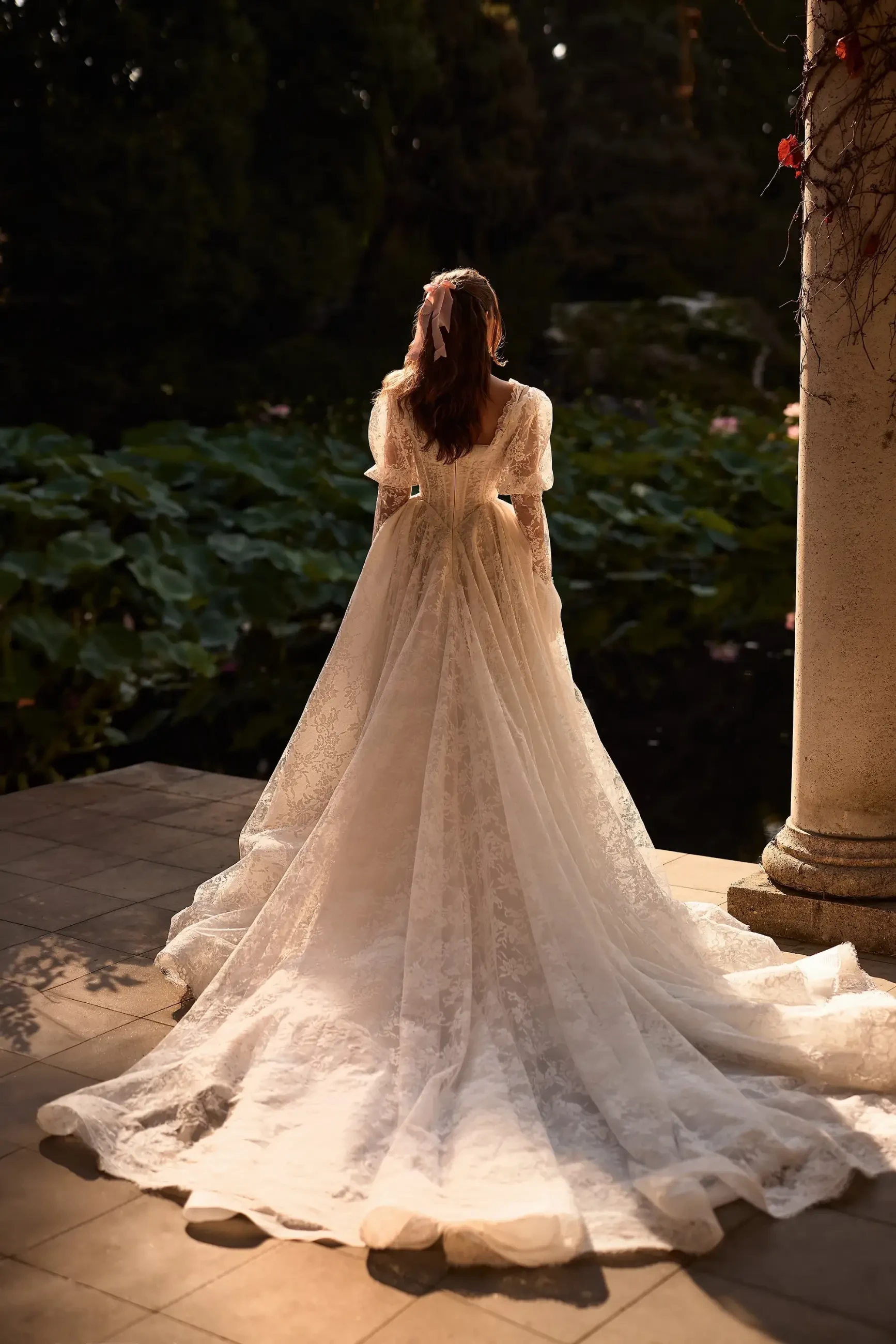 A woman stands on a stone patio, wearing a flowing, lace wedding dress with a long train, gazing at a tranquil garden pond. Soft sunlight creates a romantic, serene atmosphere.