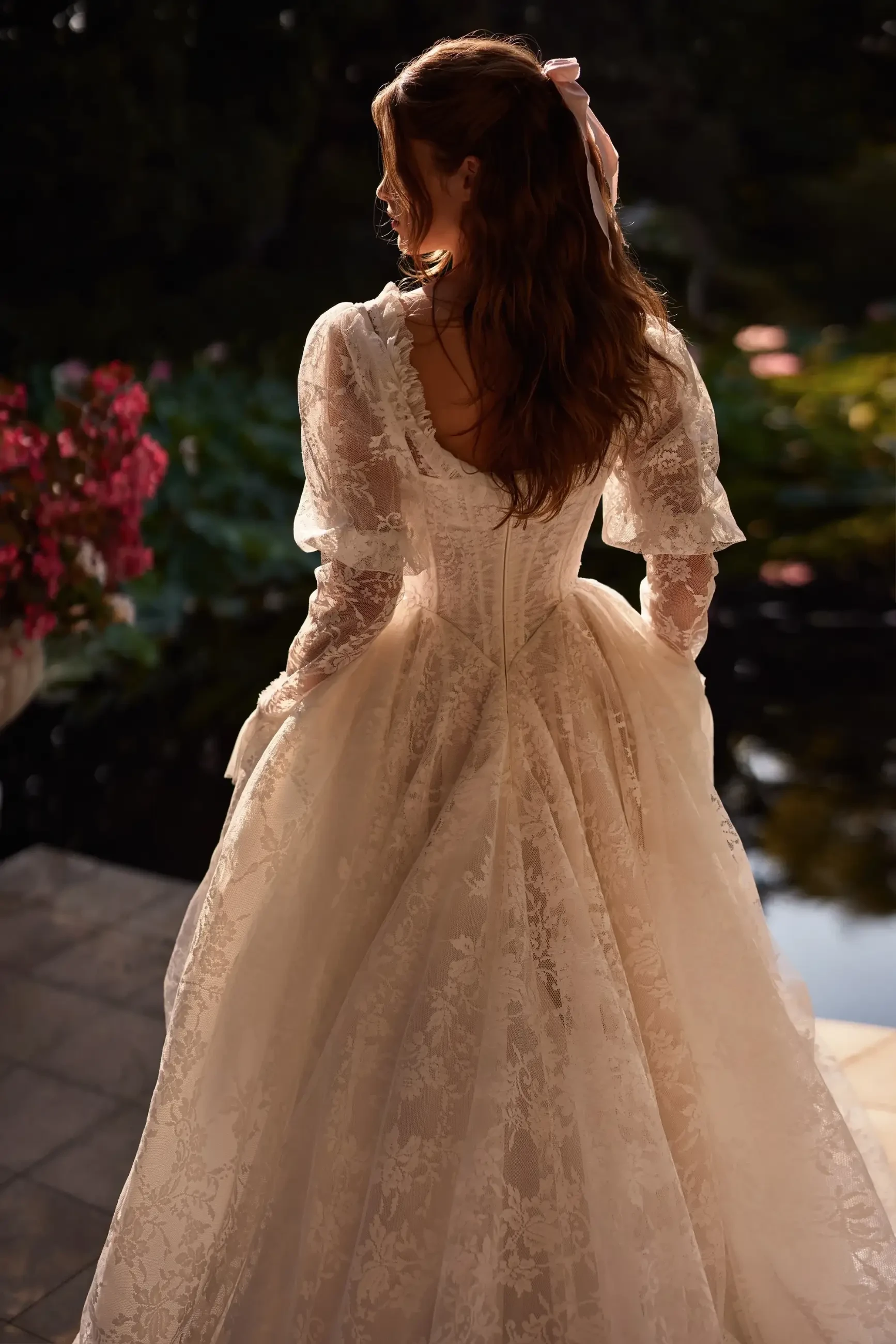 A woman in a vintage lace gown, with voluminous sleeves and a corseted back, stands outdoors. Her wavy hair features a ribbon, and pink flowers bloom nearby.