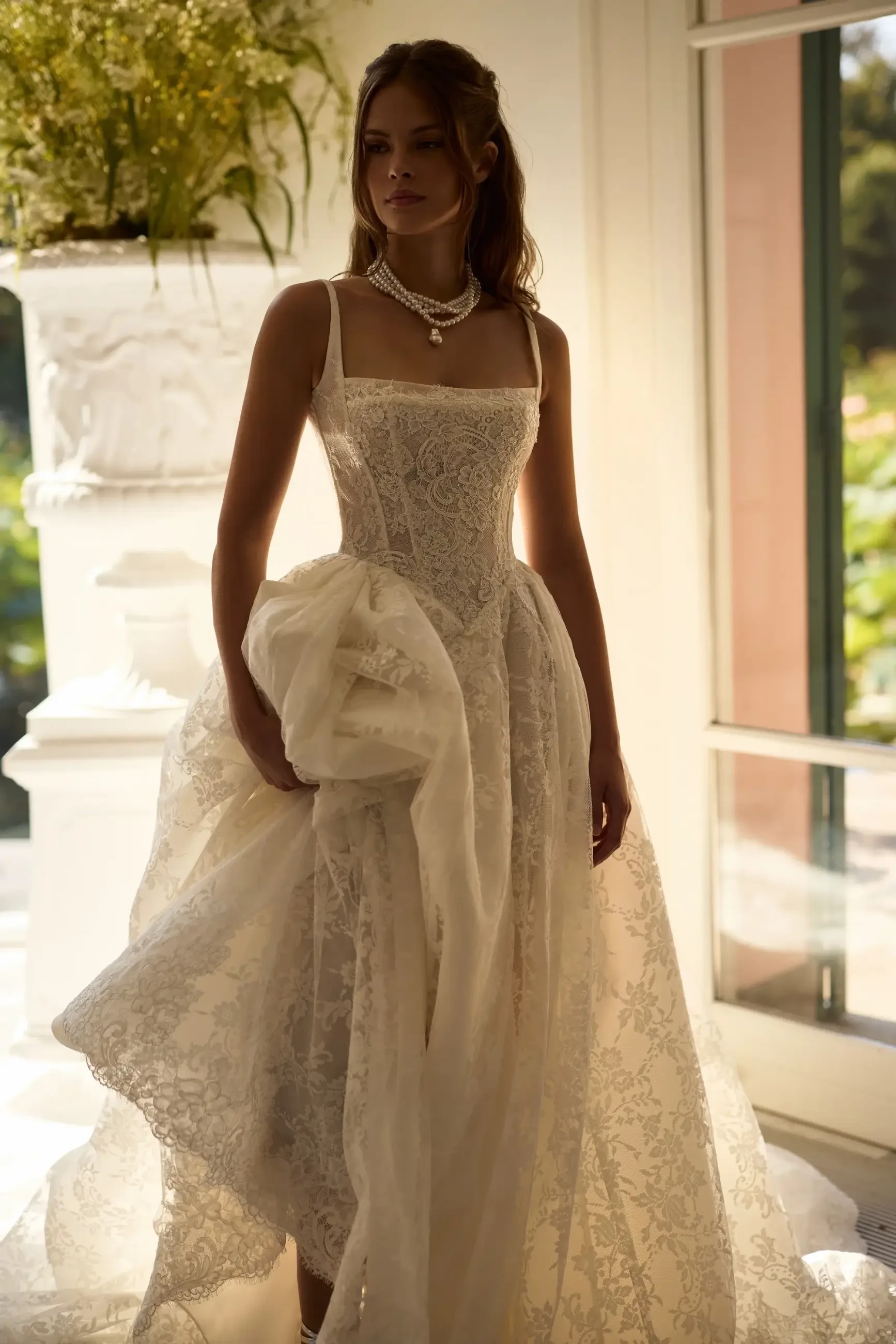 A woman stands gracefully in a sunlit room, wearing an elegant lace wedding dress. She holds the skirt slightly, exuding a peaceful, romantic aura.