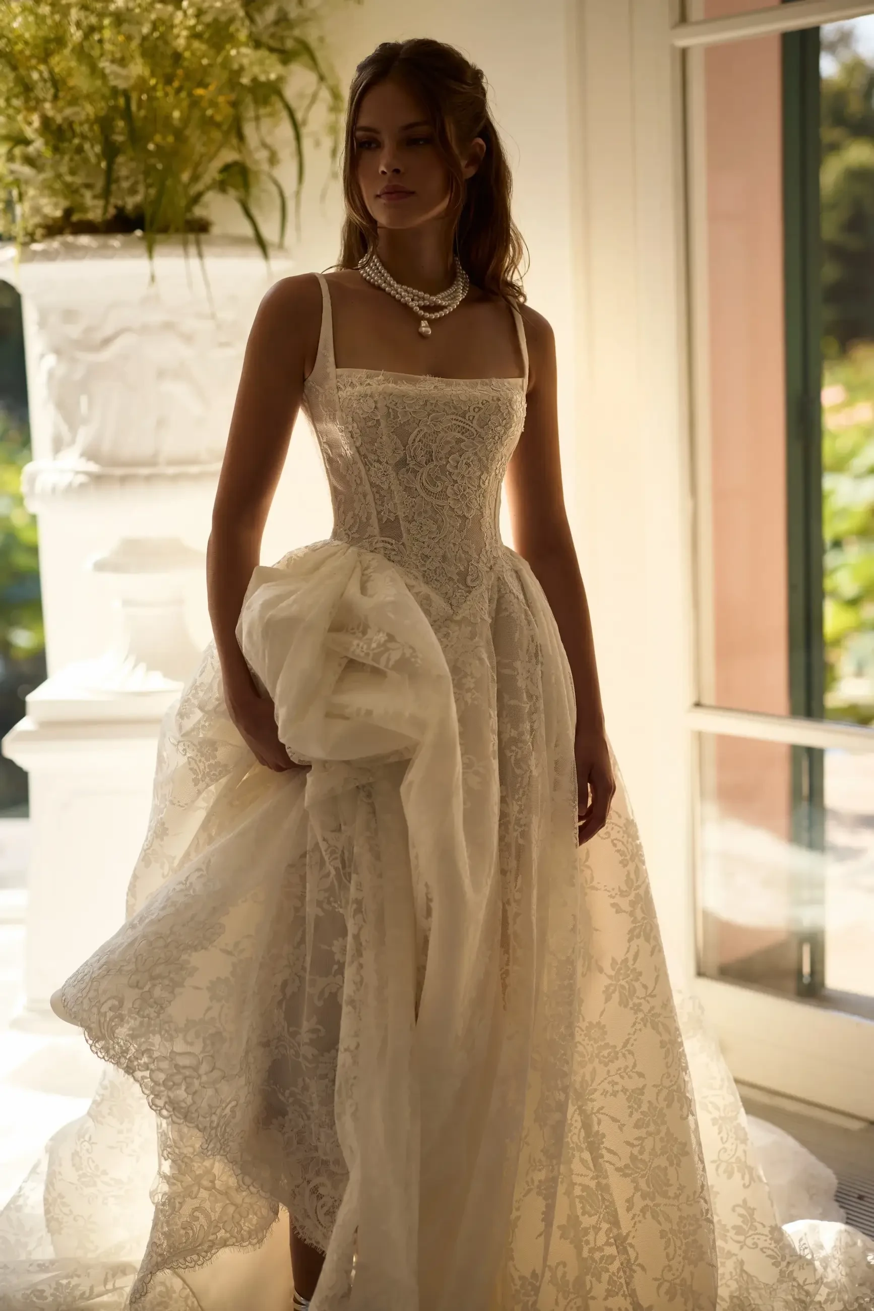 A woman wearing an elegant, intricate wedding dress with a square neckline and a flowing skirt, standing near a large window with soft natural light illuminating the scene.