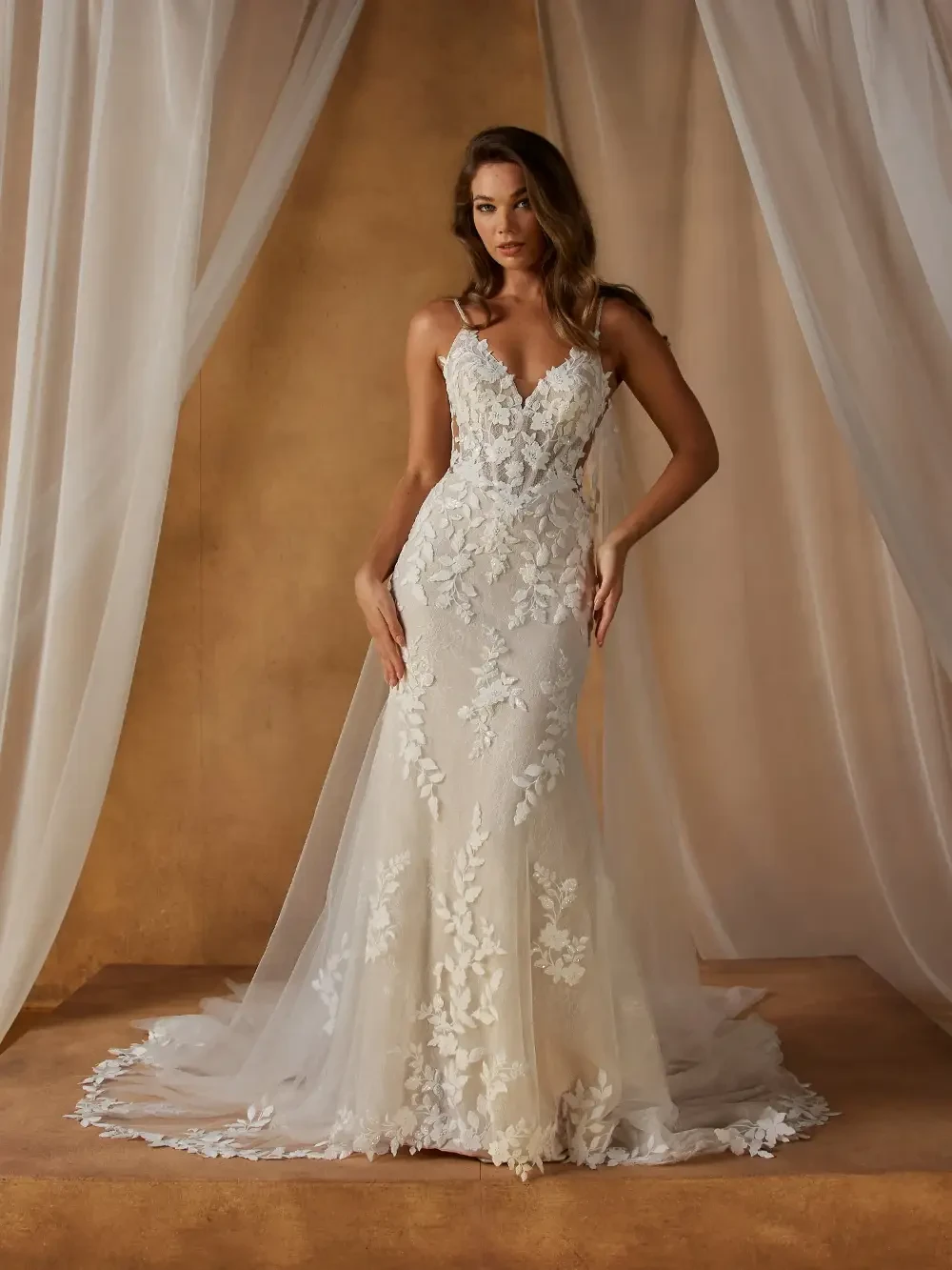 A woman in a beautiful wedding dress with intricate floral designs standing in front of a draped backdrop.