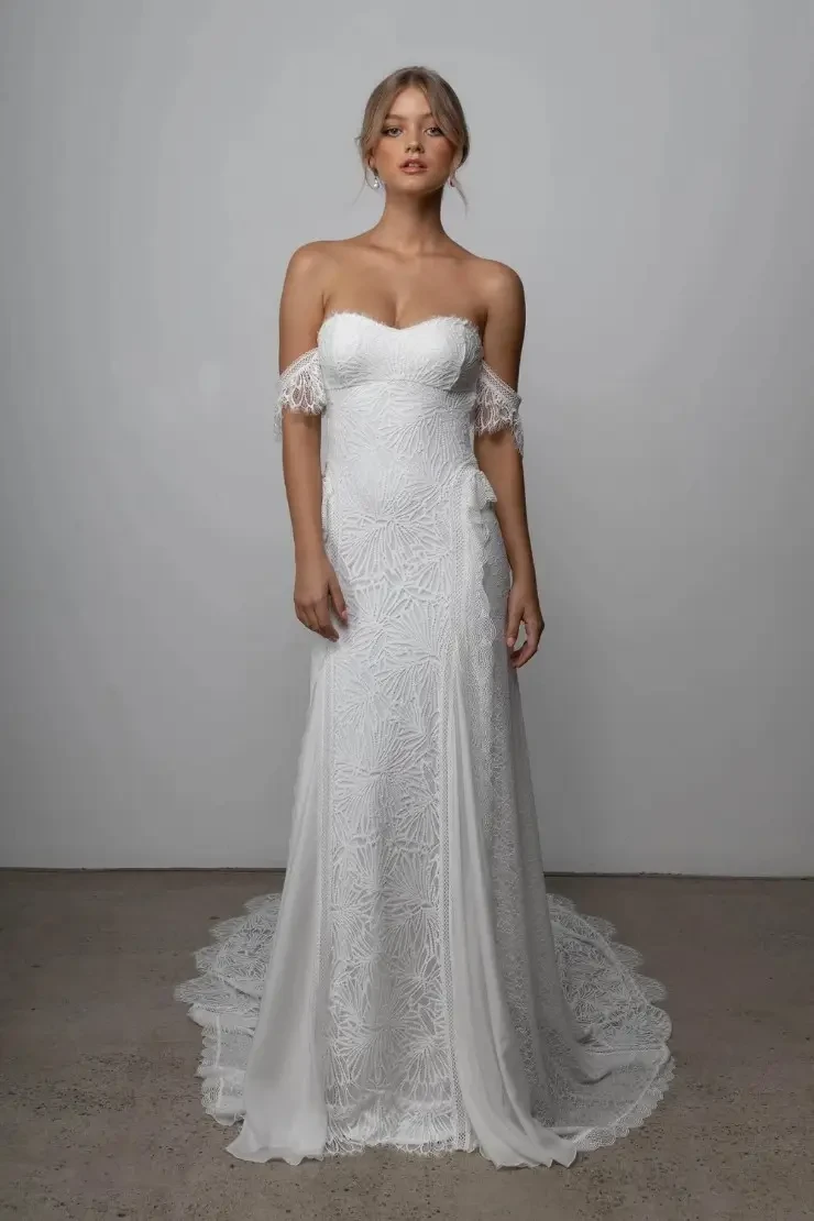 Model is wearing a feminine off-the-shoulder lace gown with scalloped armbands and back strap, crafted in stretch lace