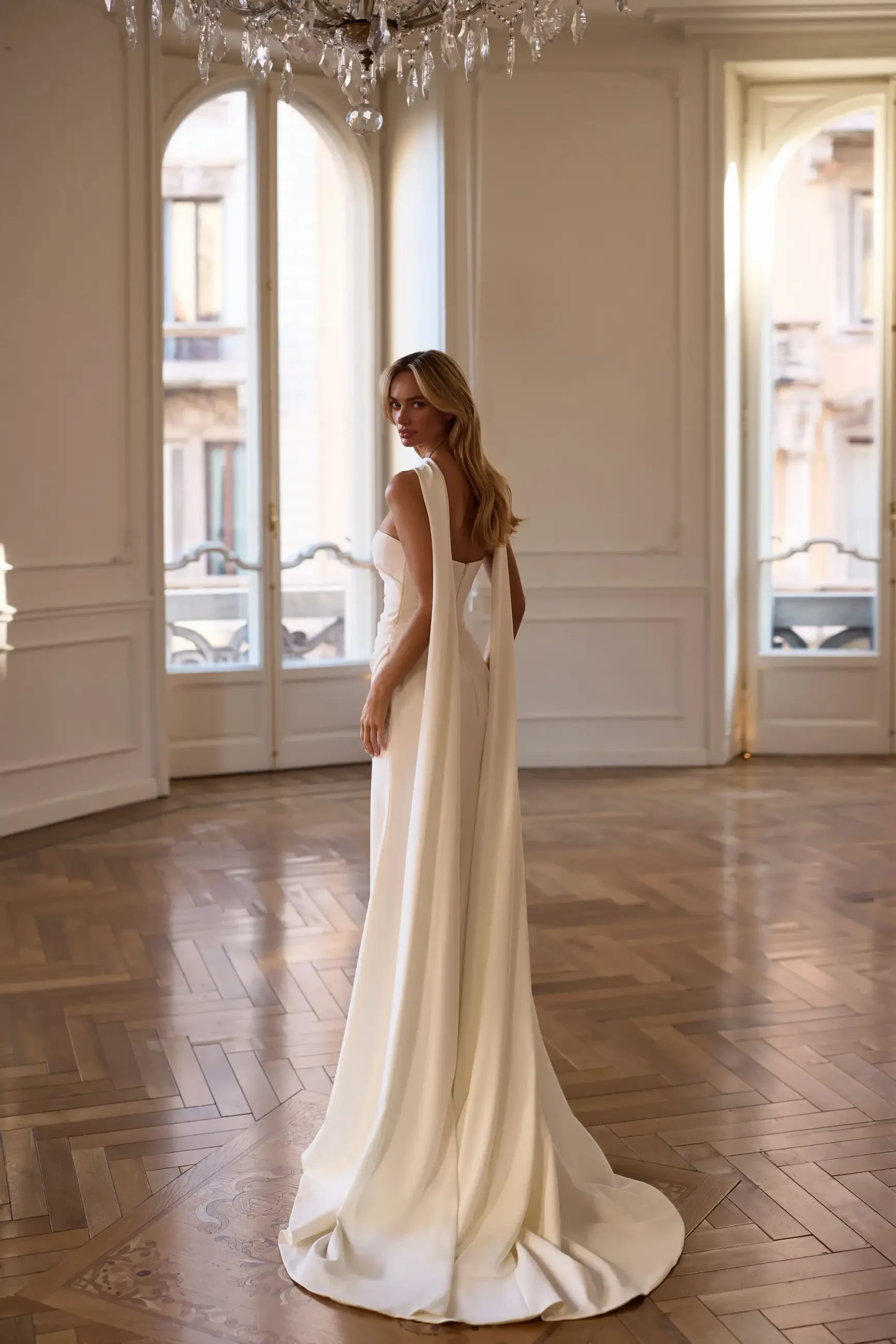 Model is wearing a minimalist strapless wedding gown with ruched detailing that contours the body.