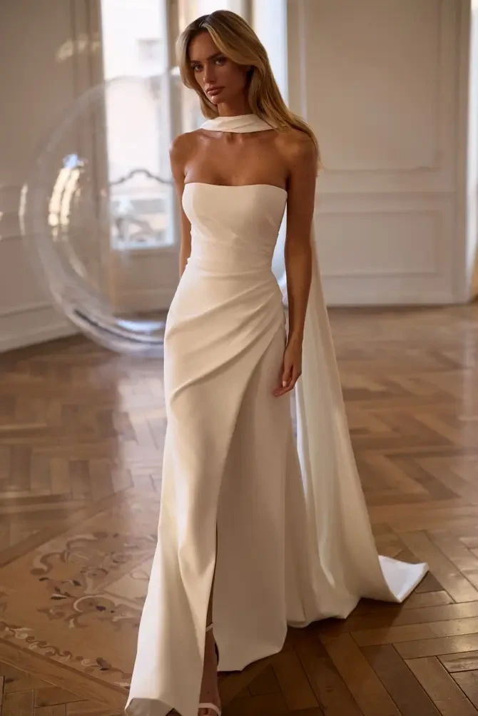 Model is wearing a minimalist strapless wedding gown with ruched detailing that contours the body.