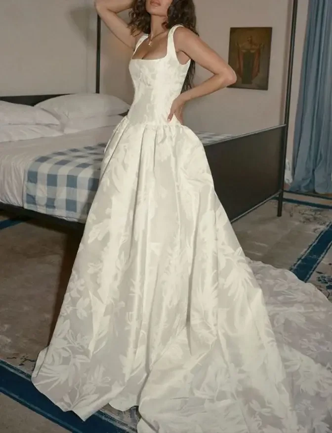 A woman wearing a white, floral-patterned wedding dress, standing in a room with a bed and a blue checkered blanket.