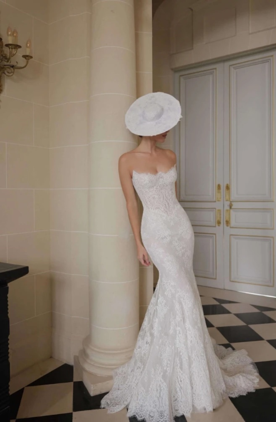 A woman in an elegant, lace mermaid-style wedding gown and wide-brimmed hat stands beside a pillar in a classic, opulent room with checkered floors.