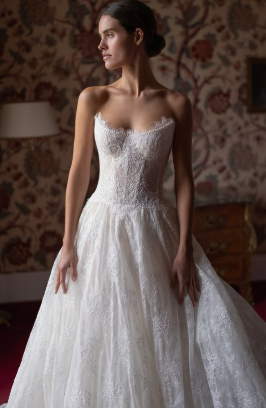 Woman in an elegant lace wedding dress with a strapless bodice, stands in a richly decorated room with floral wallpaper, exuding a serene and graceful aura.