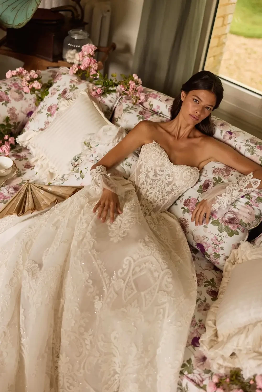 A woman in a strapless, intricately designed wedding dress lies on a floral-patterned couch, surrounded by cushions and flowers.